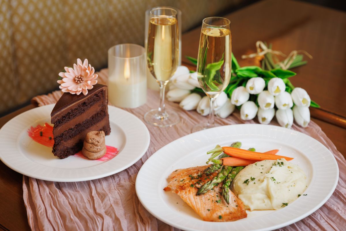 Piece of chocolate cake alongside a plate with salmon, mashed potatoes, and carrots with two glasses of champagne